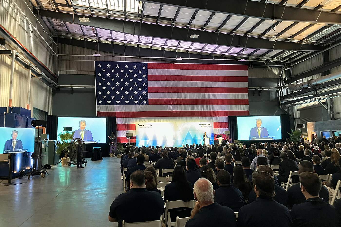 Photo: Aurubis: Several people sit on chairs in a hall, in front are large screens and the USA flag