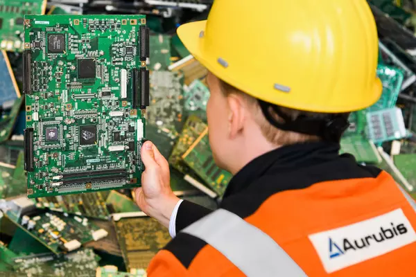 Photo: Aurubis: Man with yellow helmet and orange Aurubis clothing can be seen from behind, holding microchip plates in his hand